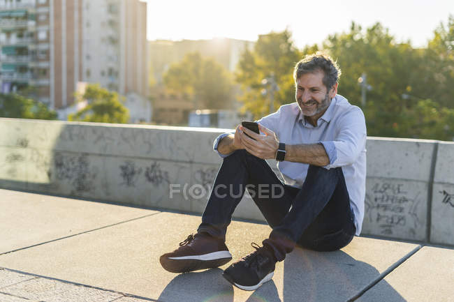 Sorridente uomo maturo seduto in città e utilizzando il telefono cellulare — Foto stock