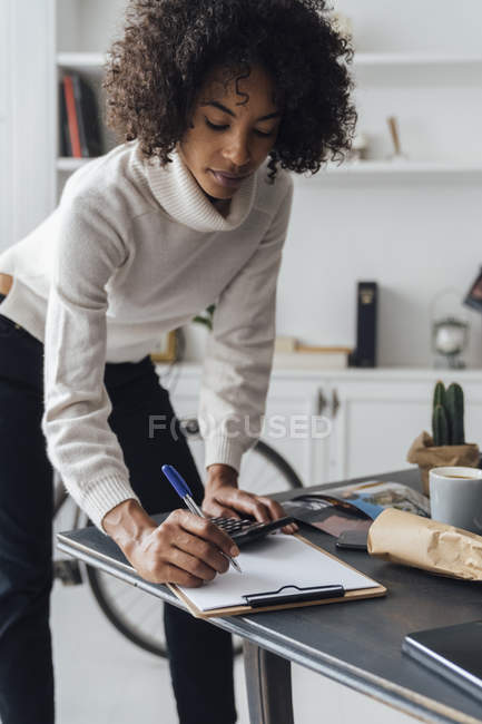 Freelancer standing at hert desk, using calculater, taking notes — Stock Photo