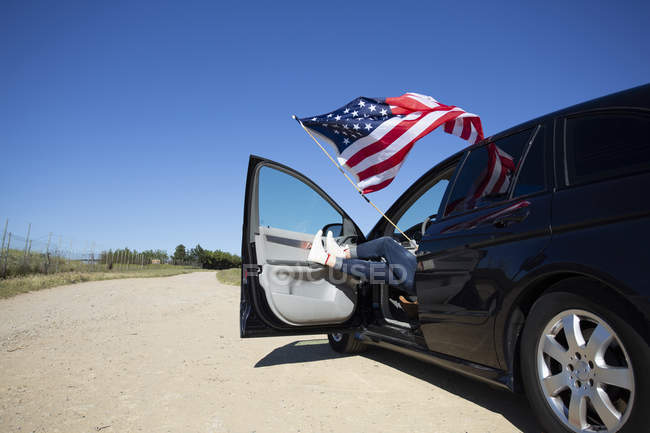 Ragazza con bandiera americana seduto in auto sul sentiero in paesaggio remoto — Foto stock