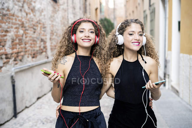 Portrait of smiling twin sisters listening music with headphones and cell phones in the city — Stock Photo