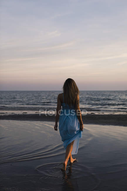 Visão traseira da jovem mulher vestindo vestido azul andando na praia ao pôr do sol — Fotografia de Stock