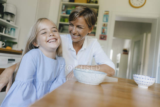 Feliz madre e hija sentadas en la mesa de la cocina en casa - foto de stock