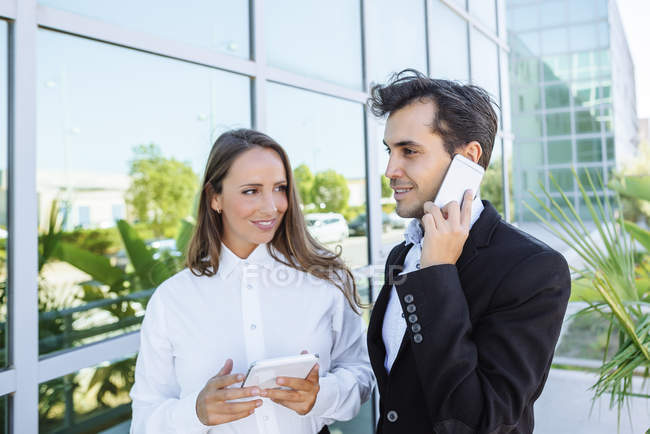 Mujer de negocios sonriente y hombre de negocios con tableta y teléfono celular fuera del edificio de oficinas - foto de stock