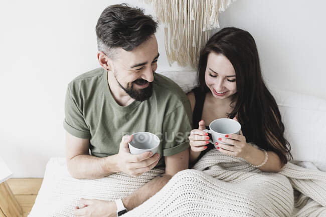 Happy couple sitting in bed, drinking coffee, having fun — Stock Photo