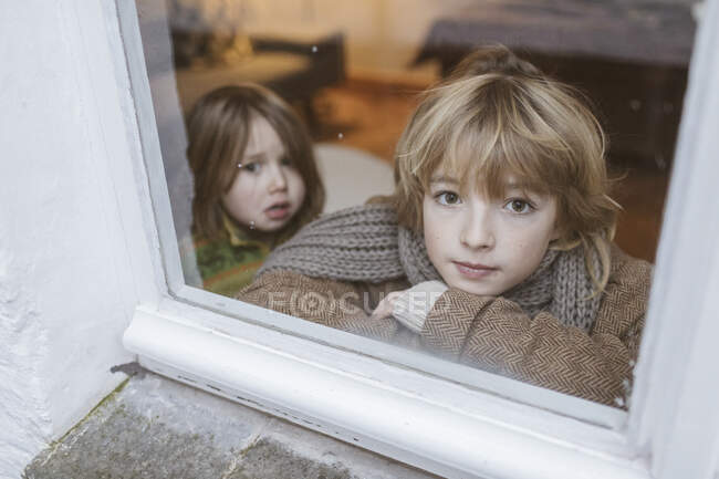 Portrait of boy looking out of window with his little sister in the background — Stock Photo