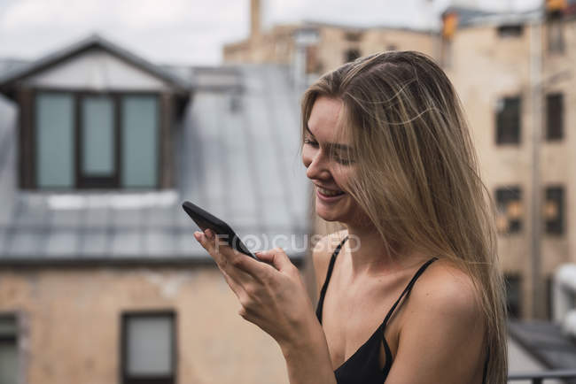 Улыбающаяся блондинка на террасе на крыше смотрит на мобильный телефон — стоковое фото