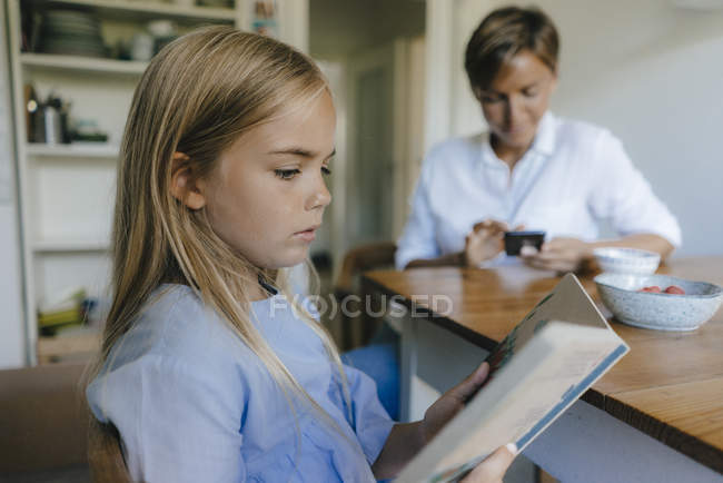 Girl with book sitting at table at home with mother using cell phone in background — Stock Photo