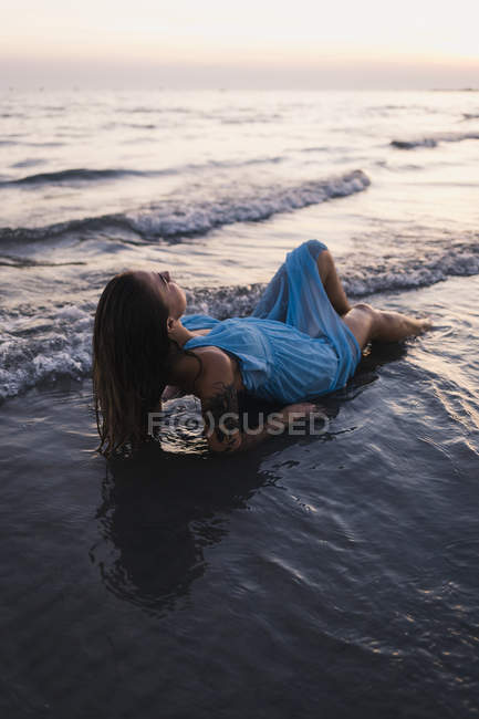 Jovem mulher tatuada vestindo vestido azul deitado na água na praia ao pôr do sol — Fotografia de Stock