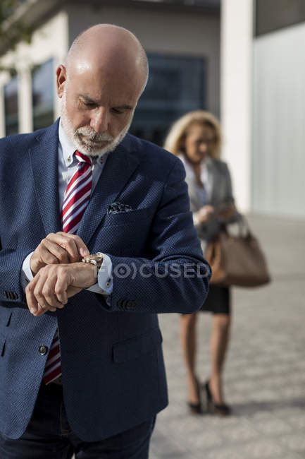 Hombre de negocios sénior comprobando el tiempo con la mujer de negocios en segundo plano - foto de stock