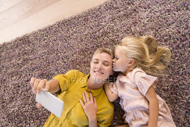Улыбающиеся мать и дочь лежат на ковре, делая селфи — стоковое фото