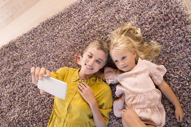 Smiling mother and daughter lying on carpet taking a selfie — Stock Photo