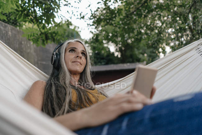 Porträt einer Frau mit Handy, die in der Hängematte im Garten liegt und mit Kopfhörern Musik hört — Stockfoto