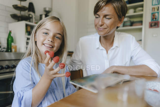 Happy mother and daughter sitting at table in kitchen at home and playing with raspberries — Stock Photo