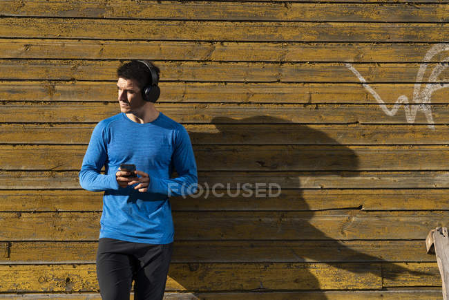 Спортсмен, що спирається на стіну будинку з мобільним телефоном та навушниками — стокове фото