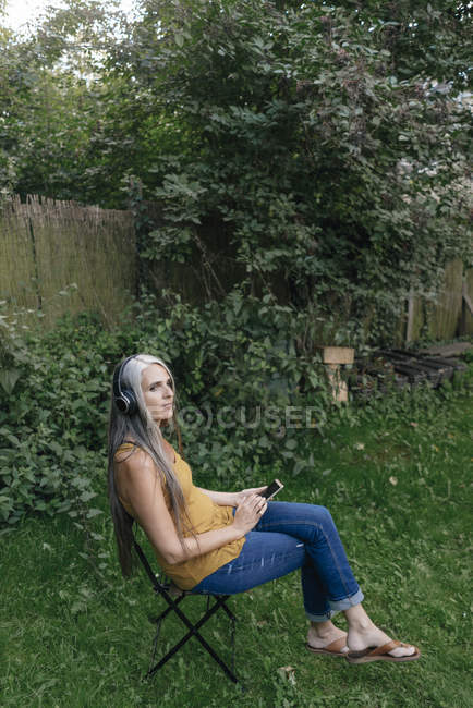 Женщина с сотовым телефоном сидит в саду и слушает музыку с наушниками — стоковое фото