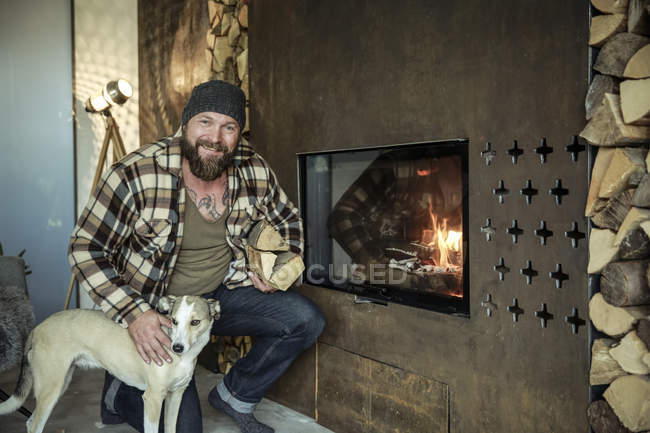 Бородатий чоловік з собакою на камін в домашніх умовах, людина тримає дрова і дивлячись на камеру — стокове фото