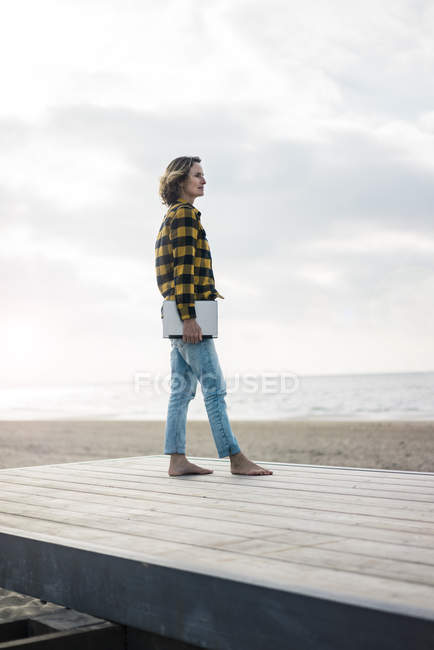 Donna matura in piedi sul lungomare in spiaggia, con in mano un computer portatile — Foto stock