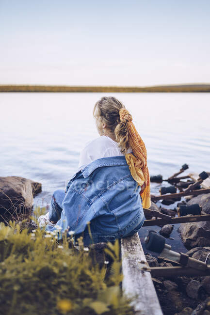 Svezia, Lapponia, vista posteriore della giovane donna seduta al bordo dell'acqua guardando la distanza — Foto stock