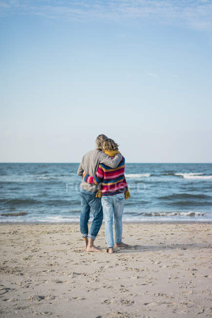 Vista posteriore di coppia in piedi sulla spiaggia con le braccia intorno, guardando il mare — Foto stock