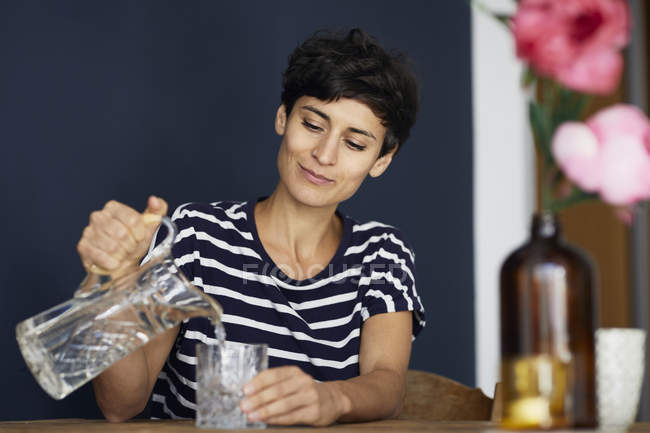 Donna a casa seduta al tavolo di legno e versare acqua nel vetro — Foto stock