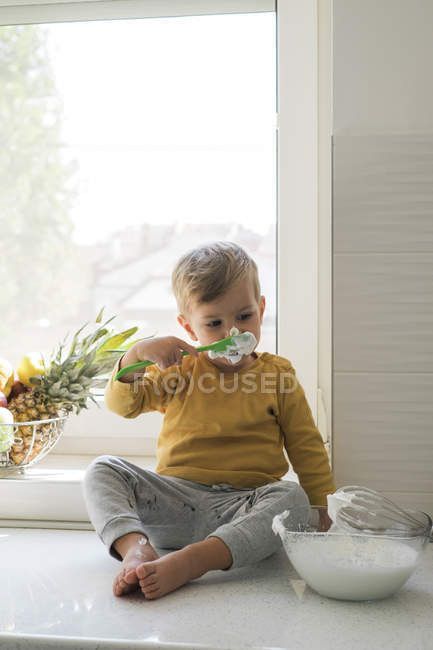Retrato de niño sentado descalzo n encimera en la cocina mordisqueando crema batida - foto de stock