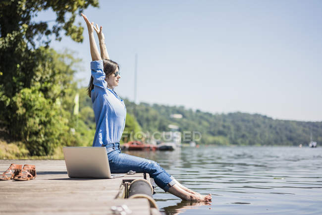 Mulher com laptop sentado em um molhe em um lago, alongamento — Fotografia de Stock