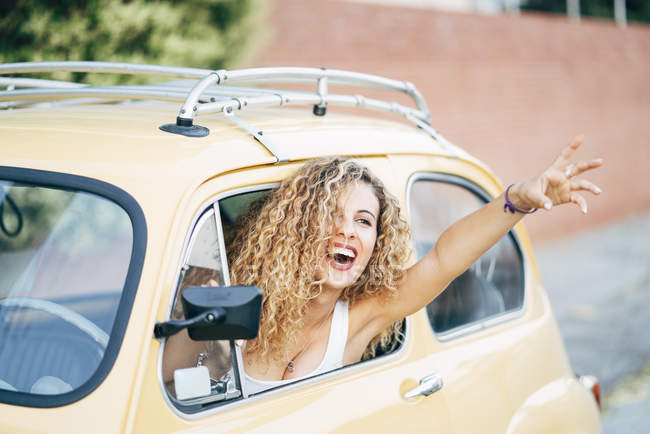 Portrait of happy blond woman leaning out of window of classic car and showing victory sign — Stock Photo