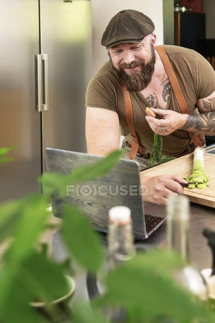 Mature man eating carrot and reading recipes in laptop — Stock Photo
