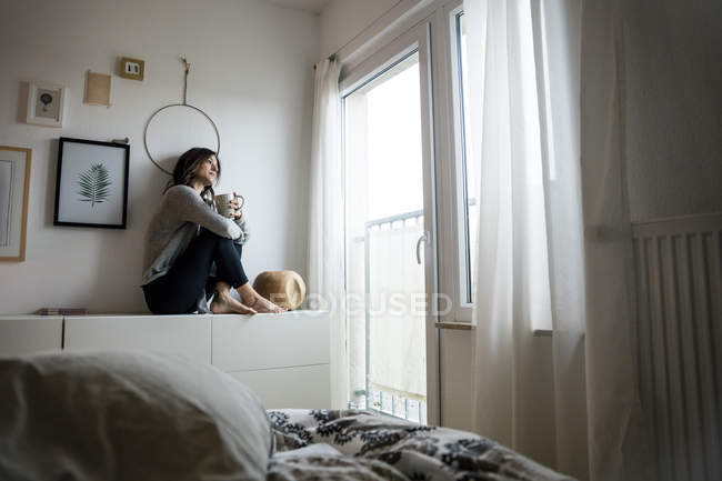 Woman sitting on chest of drawers, drinking tea, relaxing — Stock Photo