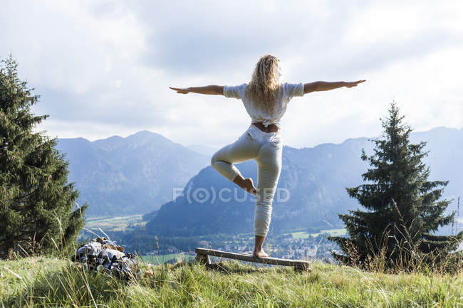 Германия, Бавария, Обераммергау, молодая женщина, занимающаяся йогой на скамейке на горном лугу — стоковое фото
