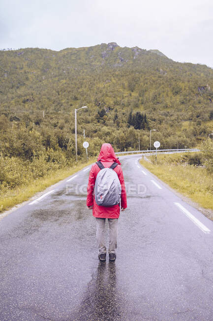 Norvegia, Lofoten, vista posteriore dell'uomo che indossa giacca pioggia rossa e zaino in piedi sulla strada bagnata — Foto stock