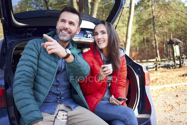 Casal apontando e olhando para longe enquanto sentado no tronco do carro durante viagem de estrada — Fotografia de Stock