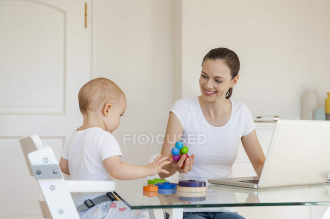 Sorrindo mãe usando laptop brincando com a filhinha na mesa em casa — Fotografia de Stock