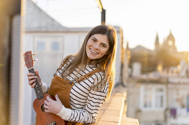 Ritratto di giovane donna sorridente in piedi sulla terrazza sul tetto la sera giocando ukulele — Foto stock