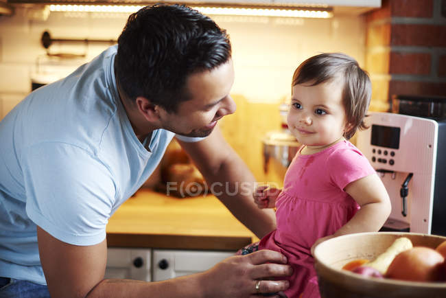 Smiling father looking at baby girl sitting on counter in kitchen at home — Stock Photo
