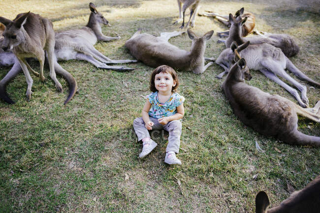 Australien, Brisbane, Porträt eines lächelnden kleinen Mädchens, das auf einer Wiese zwischen zahmen Kängurus sitzt — Stockfoto
