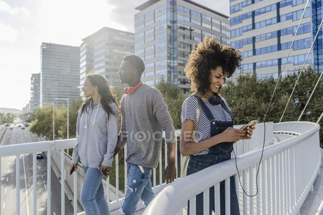 Молодая женщина в наушниках, стоящая на мосту, используя смартфон, молодые люди проходят мимо — стоковое фото
