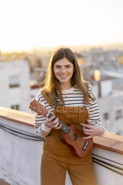 Retrato de sorrindo jovem mulher de pé no terraço do telhado à noite jogando ukulele — Fotografia de Stock