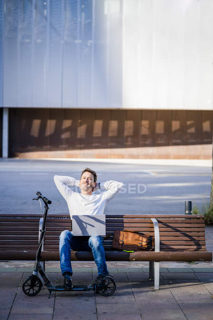 Casual businessman with kick scooter, sitting on a bench, working relaxed in the city — Stock Photo