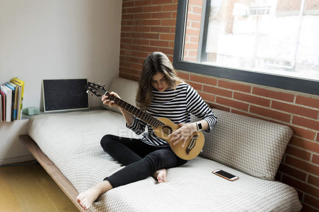 Mujer joven sentada en el sofá en casa y tocando la guitarra - foto de stock