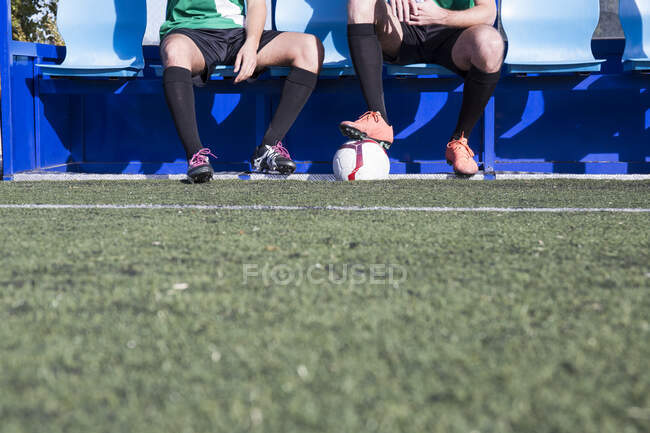 Ноги двух футболистов сидят на скамейке на футбольном поле — стоковое фото