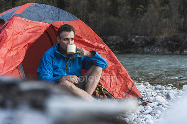 Hombre maduro acampando a orillas del río, bebiendo - foto de stock