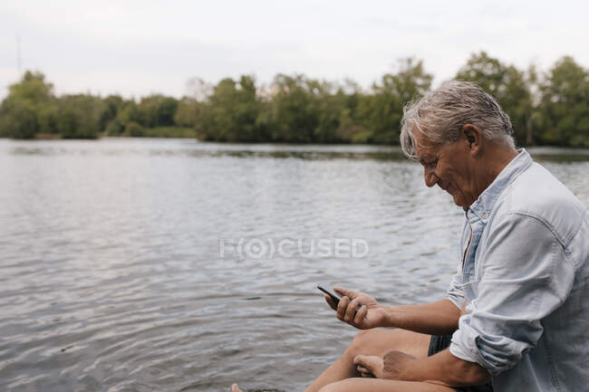 Senior man sitting at a lake using cell phone — Stock Photo