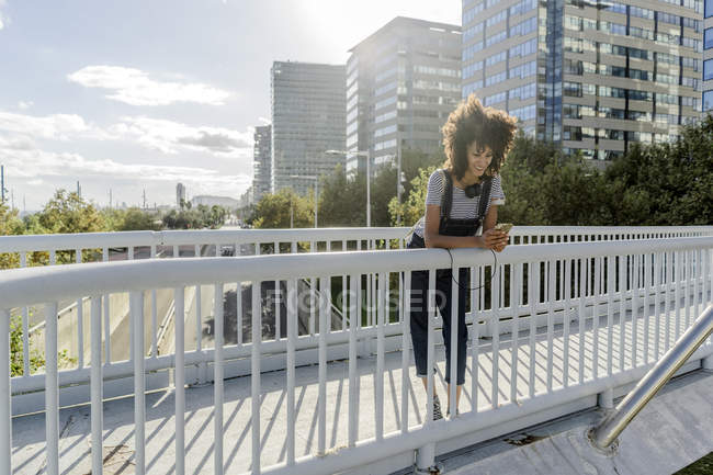 Jovem de pé em uma ponte, usando smartphone, com fones de ouvido ao redor do pescoço — Fotografia de Stock