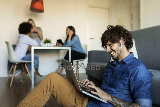 Un homme souriant assis par terre sur un ordinateur portatif avec des amis en arrière-plan — Photo de stock