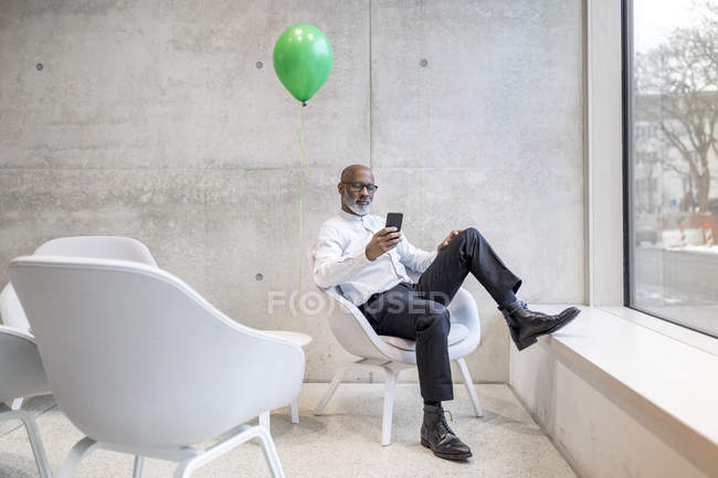 Зрелый бизнесмен с зеленым шаром сидит на кресле и смотрит на мобильный телефон. — стоковое фото