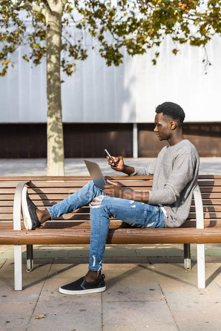 Joven sentado en un banco de la ciudad, usando un ordenador portátil y un teléfono inteligente - foto de stock