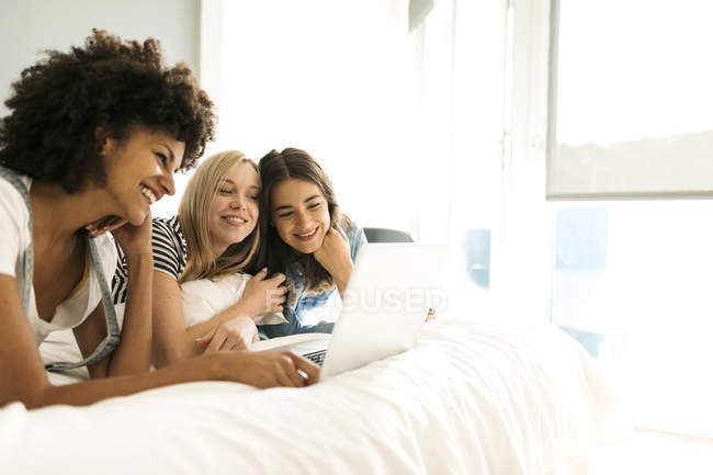 Три счастливые подруги, лежащие на кровати, делят ноутбук — стоковое фото