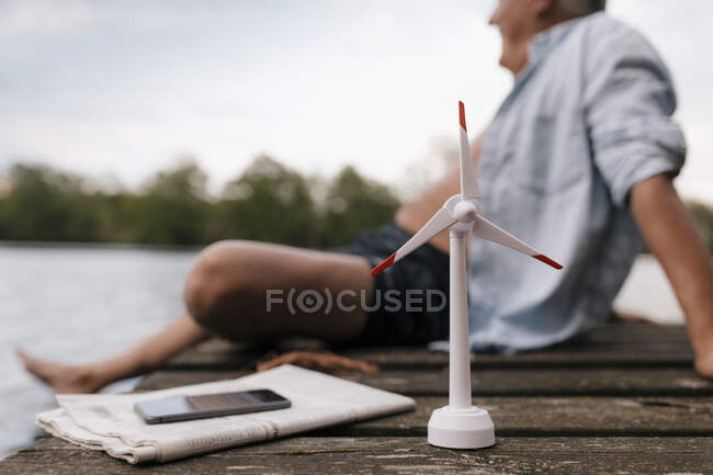 Senior man sitting on jetty at a lake with small wind turbine model. - foto de stock