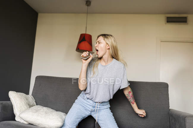 Jeune femme exubérante sur un canapé avec téléphone cellulaire faisant semblant de chanter — Photo de stock
